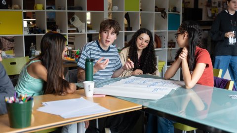A group of diverse young adults collaborates around a table with notes and materials, engaged in discussion in a creative workspace.