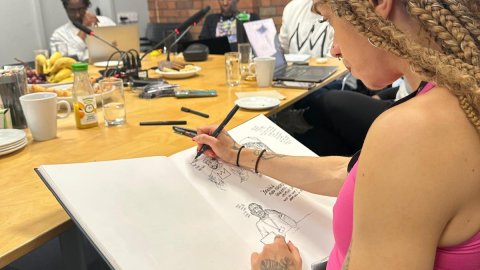 A person with braided hair sketches in a large notebook during a meeting at a wooden table with food and drinks present.