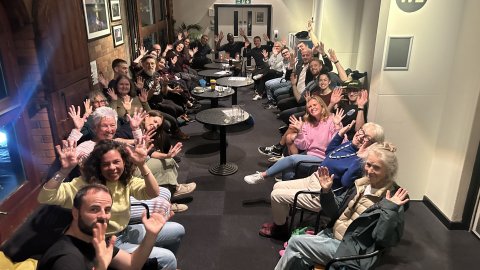 hoto of Deaf Conversations About Cinema discussion group at Watershed. A large group of people sitting in a long corridor together - waving their hands in the air after the discussion.