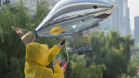 Person in a yellow raincoat holding a large silver shark-shaped balloon near a river with trees and city buildings in the background.