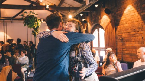 Two people hug and smile in a room with exposed brick walls, while others sit and watch in the background.
