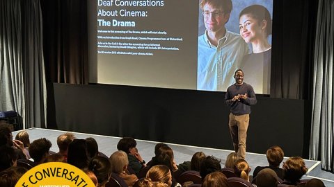 A man stands in front of a large cinema screen talking to an audience in the cinema