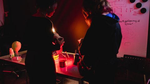 Two people examine a small object at a cluttered worktable under red light, beside a whiteboard covered in sketches and notes; the mood is focused and tense.