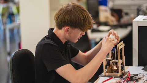 A focused young man in a black polo carefully assembles an electronics project at a workbench, surrounded by wires and tools in a busy workshop.