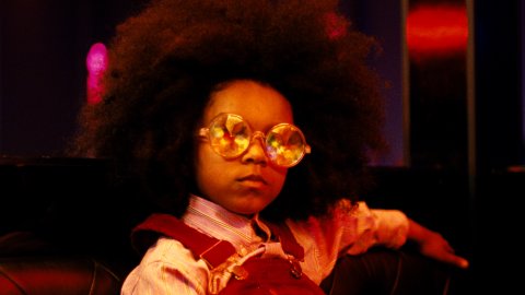 A child wears large, round mirrored sunglasses and dungarees and looks towards the camera. 