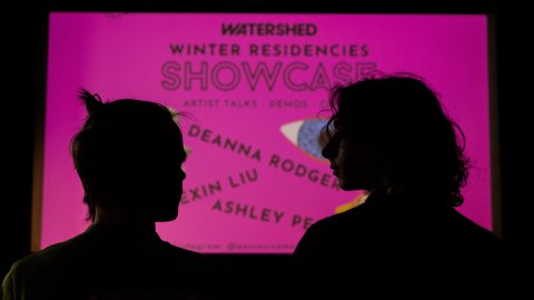 Silhouettes of two figures from the shoulders up. Before them is a neon pink sign, reading "Watershed Winter Residencies Showcase".