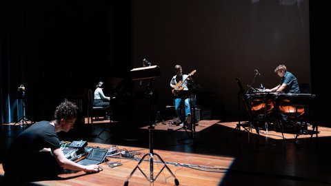 A group of musicians in a dark studio. One plays guitar, one drums and a piano in the background.