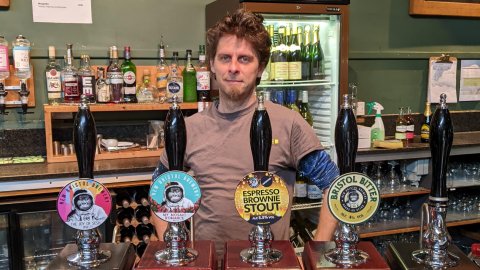 An image of a man standing behind four beer pumps.