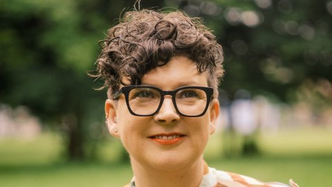 Head and shoulders of a woman with short hair and glasses wearing a multi coloured, stripey top with trees in background.