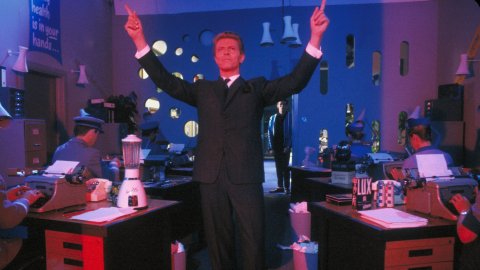 Daivd Bowie pointing his hands in the air standing amongst desks.