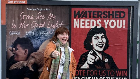 An outdoor poster site - a person stands to the side smiling and pointing in the same pose as the person in the poster which also has the words' Watershed Nees