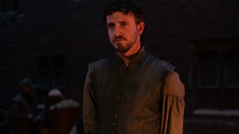 A young man stands in dim light, wearing a historical tunic with an embroidered collar, while figures gather in the background.