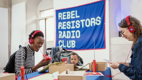 A young Black women sits smiling with red headphones on and a banner above her reads Rebel Resistors Radio Club. 