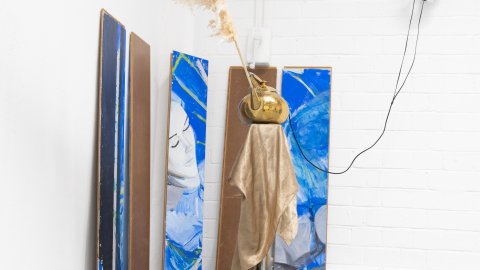 A minimalistic setup features headphones on a wall mount, a framed artwork above a yellow cloth-draped shelf, and two brown vases with pampas grass below.