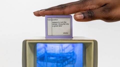 A hand hovers over a small screen atop a retro TV showing a blue image. The screen displays text with words about a "palace" and a "lake." The vibe is artistic and thought-provoking.