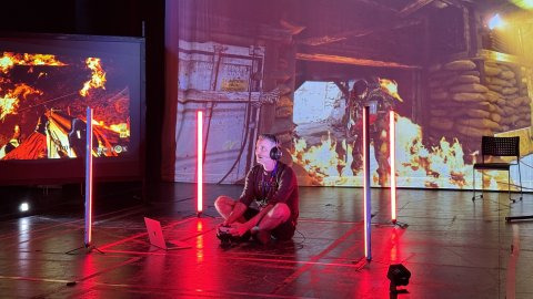 A person sits cross-legged on the floor, wearing headphones, surrounded by vertical red lights. Intense video game scenes with fire project in the background.
