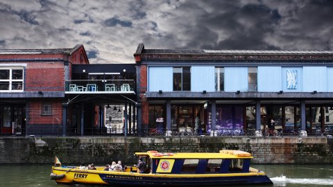 exterior shot of the Watershed building, including the balcony, there is a yellow ferry on the water passing the building and the sky is dark and stormy.