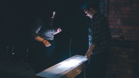 Two people stand in a dimly lit room, focused on a glowing rectangular table. The scene conveys curiosity and concentration against a brick wall backdrop.