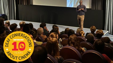 Inside a full cinema, man at the front of the audience introduces a Descriptive Subtitled screening of The Drama