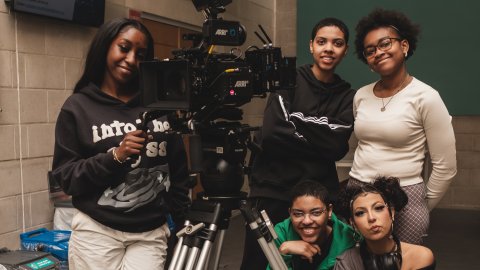 Five young people posing with a professional ARRI camera on a tripod in an indoor setting with concrete walls.