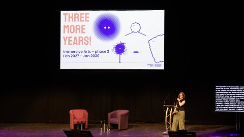Speaker stands on a dimly lit stage beside a slide reading “THREE MORE YEARS!” with colorful drawings, creating an optimistic, creative tone.