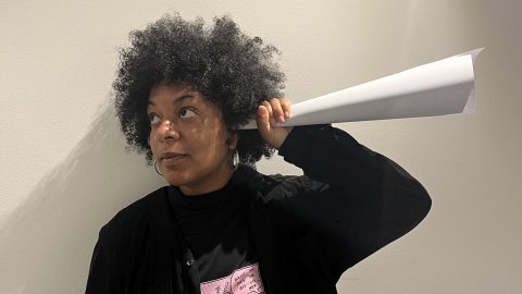 A photo of a woman holding a paper cone up to her ear as if she is listening to it.