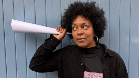 A photo of a woman holding a paper cone up to her ear as if she is listening to it.
