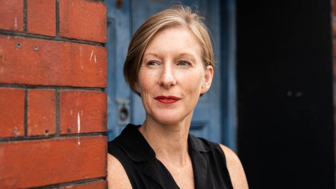 A woman with short hair leans against a brick wall with a blue background, she is smiling and looking to here right.