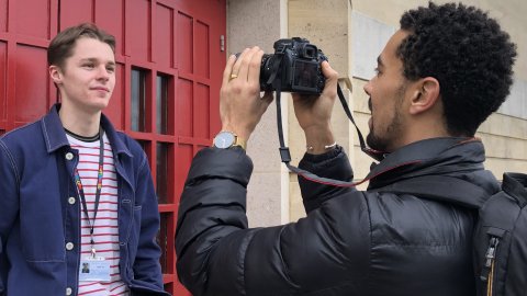 One man taking a photograph of another man who is standing smiling in front of a red door.