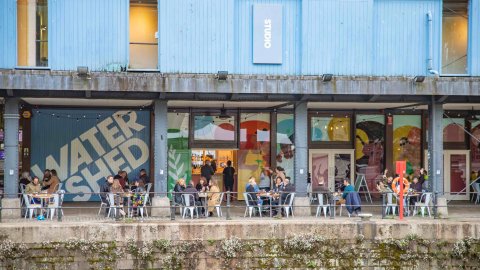 An image of people sitting at tables outside of Watershed's blue sheds, overlooking the harbour.