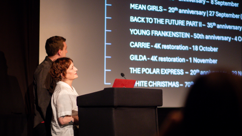 Two people are presenting in front of a cinema screen.