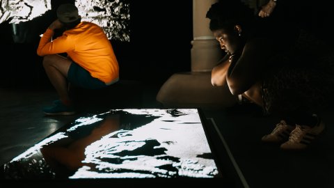 A young Black person is peering over an illuminated screen of FRAMERATE PULSE OF THE EARTH.