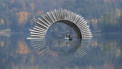The scene is slightly misty, a man sits in a blue boat with oars on open water inside a sculpture. There are autumn trees in the background.