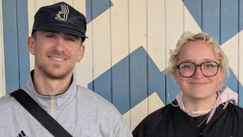A young man in a grey jacket and cap stands next to a woman with short, wavy blonde hair, in front of a blue and white patterned wall.