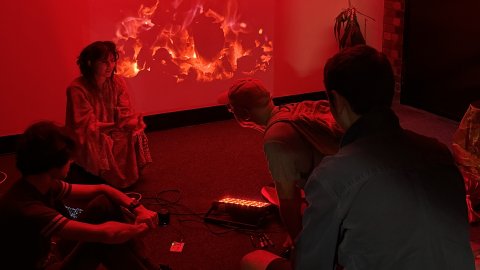 A group of people sit in a dimly lit room with red lighting, focused on a vibrant fire projection on the wall. Modern art hangs above them.