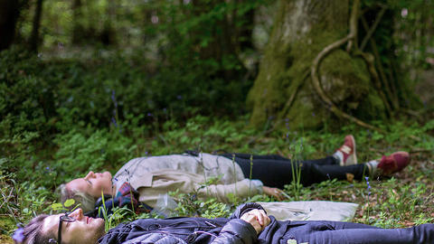 Still from Afterlife - two people lying in a wood listening to headphones
