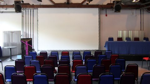 Photo of Waterside 3 at Watershed – a large room with chairs facing a projector screen, a speakers podium and top table