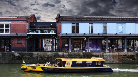A photo of the Watershed building, on Bristol old docks with a passenger ferry passing by.