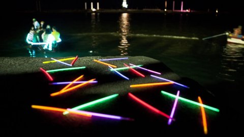 An image of a boat on the water in the distance at night with glow sticks laid out on the pavement at the front of the image which say 'lets play'.