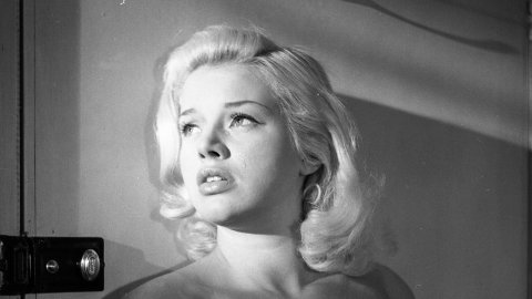 A black and white film still. A woman stands with her back against the door, looking troubled.