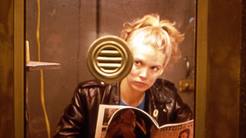 A woman in a glass-fronted booth. She reads a magazine with topless women on the cover.