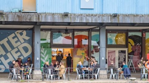 An image of people sitting on chairs having a drink at Undershed bar in front of the Watershed sign.
