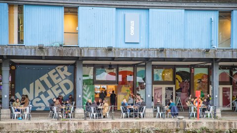 The exterior of Watershed, a victorian dockside warehouse with, people sitting on café chairs and a large Watershed log on the from o the building