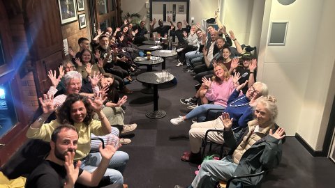A diverse group of people sitting in a cozy, dimly lit venue, all raising their hands enthusiastically in a communal gathering.