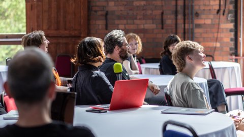 A group of attendees sits attentively in a spacious room, with tables covered in white linens and large windows letting in natural light.