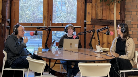 Three individuals sit at a round table in a cozy studio, equipped with microphones and laptops, surrounded by plants and large windows.