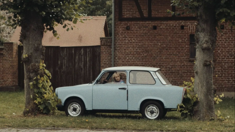 A vintage light blue car parked on grass, surrounded by trees and a rustic brick building in the background.
