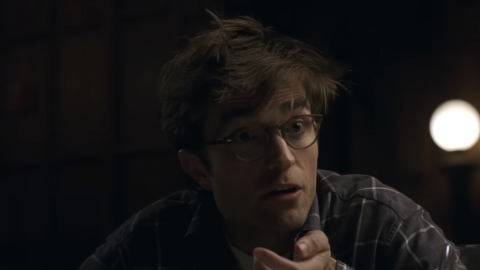 A person gestures while sitting at a table in a dimly lit room, with a warm light source in the background.