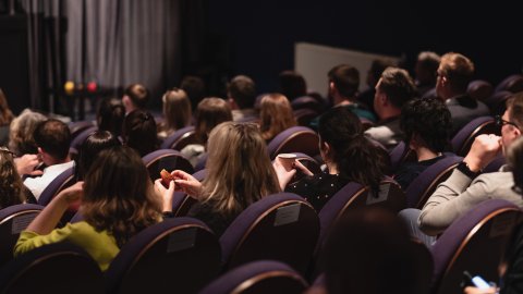 Audience seated in a dimly lit cinema, some holding snacks and drinks, facing away from the camera toward a stage.
