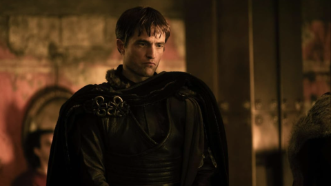 A person dressed in dark armour and cloak stands in a dimly lit room with stone walls and warm lighting.